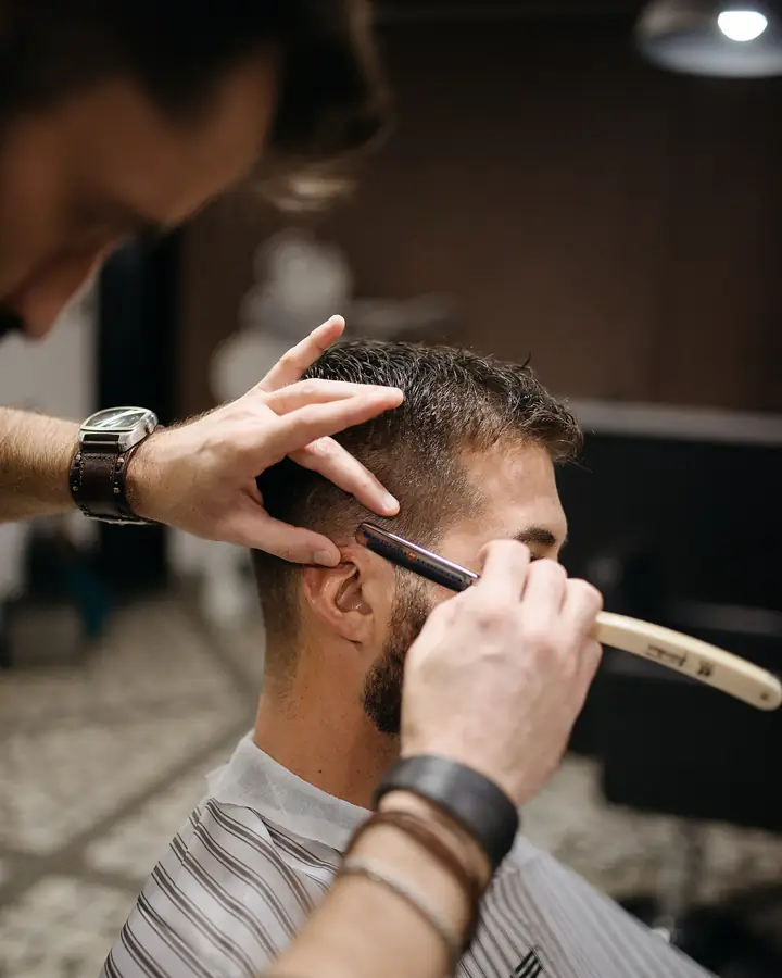 Barbero afeitando con navaja la mejilla de un cliente, en penumbra